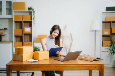 Startup small business SME, Entrepreneur owner woman using smartphone or tablet taking receive and checking online purchase shopping order to preparing pack product box.