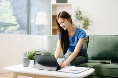 Genç Asyalı kadın evinde rahat bir kanepede rahat rahat oturup akıllı telefon ve tablet gülümseyen kızla mesajlaşırken evden cep telefonu alışverişini kullanıyor.