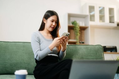 Genç Asyalı kadın evdeki kanepede akıllı telefon ve dizüstü bilgisayar kullanıyor.