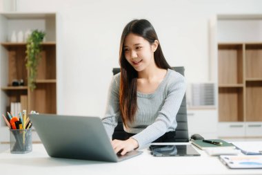 Masada oturan Asyalı bir kadın dizüstü bilgisayar kullanarak ofiste Finans ve Pazarlama ile uğraşıyor.