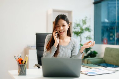 Masada oturan Asyalı bir kadın dizüstü bilgisayar kullanarak ofiste Finans ve Pazarlama ile uğraşıyor.