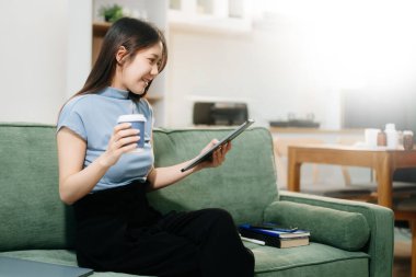 Genç Asyalı kadın evinde rahat bir kanepede rahat rahat oturup akıllı telefondan ve tabletten mesajlaşarak gülümseyen kız evden cep telefonu alışverişini kullanıyor.
