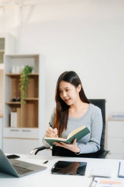 Genç çekici Asyalı kadın gülümsüyor. Not defteri, tablet ve dizüstü bilgisayarla yazmayı düşünüyor. Evden çalışıyor, ofisteki kameraya bakıyor.