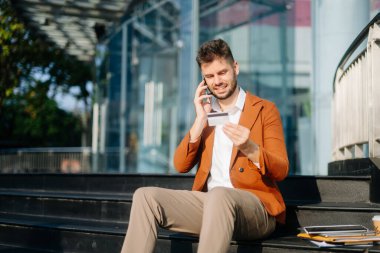 Modern iş adamı dışarıda, akıllı telefonla konuşuyor ve ofis binasının merdivenlerinde otururken kredi kartı tutuyor. 
