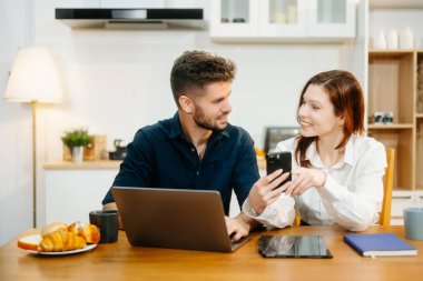 Modern bir çift, rahat bir mutfak ortamında dizüstü bilgisayar, defter ve kahvaltıyla birlikte kahve içerken iş birliği yapıyor. Yaşam tarzı, takım çalışması ve günlük verimliliğin mükemmel bir karışımı.