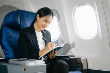Çekici Asyalı kadın uçak yolcusu dizüstü bilgisayar ve tabletle kablosuz bağlantı kullanarak rahat koltukta oturuyor. Stil içinde seyahat et, zarafetle çalış..