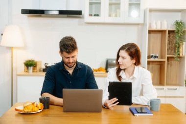 Modern bir çift, rahat bir mutfak ortamında dizüstü bilgisayar, defter ve kahvaltıyla birlikte kahve içerken iş birliği yapıyor. Yaşam tarzı, takım çalışması ve günlük verimliliğin mükemmel bir karışımı.