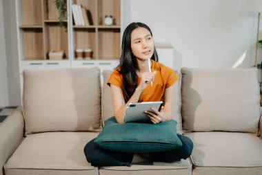 Genç Asyalı kadın evinde rahat bir kanepede rahat rahat oturup akıllı telefon ve tablet gülümseyen kızla mesajlaşırken evden cep telefonu alışverişini kullanıyor.