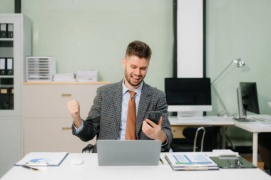 Genç işadamı akıllı telefon kullanıyor, dizüstü bilgisayarda çalışıyor ve ofiste el kol hareketi yapıyor..