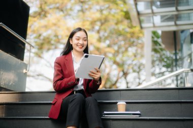 Ofisin dışında elinde not defteri ve tabletle gülümseyen kendine güvenen Asyalı kadın. İş merkezi arka planında büyük şehir