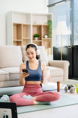 Genç bir kadın yoga yapıyor ve paspas ve jimnastik malzemeleriyle esniyor, sağlığını, motivasyonunu ve modern bir ev ortamında sağlıklı ve aktif bir yaşam tarzını gösteriyor.