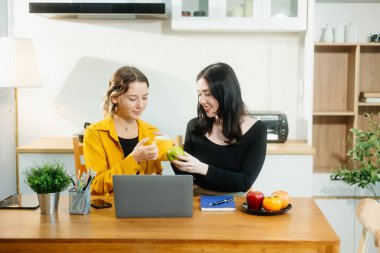 İki beyaz kadın mutfak masasında oturmuş laptoplarıyla evde elma yiyorlar. 