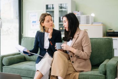 İki iş kadını, profesyonel bir ofiste koçun üzerinde oturan bir klasörle strateji kuruyor. Bu modern işbirliği konseptiyle takım çalışmasını, liderliği ve yeniliği vurgula.