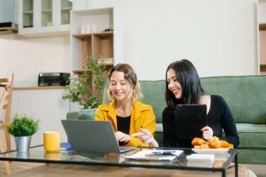 Yeşil kanepenin yanında oturan iki neşeli kadın şık bir evde dizüstü bilgisayar ve tablet kullanarak fikir alışverişinde bulunuyorlar. Takım çalışması çalışma alanı temaları için mükemmel
