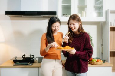 İki genç Asyalı kadın sıcacık bir mutfakta kahvaltıdan keyif alıyor, gülümsüyor, sohbet ediyor ve sıcak, rahat bir atmosferde yemek paylaşıyorlar.. 