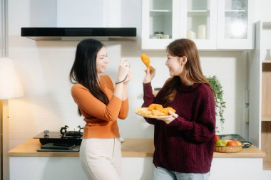 İki genç Asyalı kadın, sıcacık bir mutfakta kahvaltıdan zevk alıyorlar. Biri diğerinin sıcak ve rahat bir atmosferde fotoğrafını çekiyor.. 