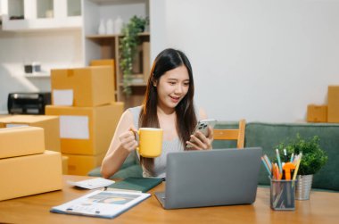 Startup small business SME, Entrepreneur owner woman using smartphone or tablet taking receive and checking online purchase shopping order to preparing pack product box. 