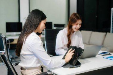 Asyalı iş arkadaşları sunar ve açıklar ve dizüstü bilgisayar ve tablet kullanır. Takım çalışması, finansal pazarlama ekibi, modern ofis odasında otururken.