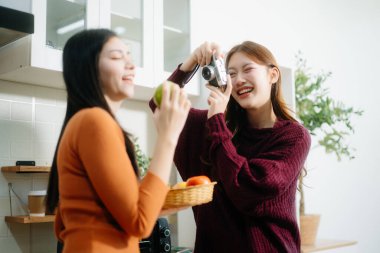 Sıcak bir mutfakta elma yiyen iki genç Asyalı kadın, biri diğerinin sıcak, rahat bir atmosferde fotoğrafını çekiyor..  