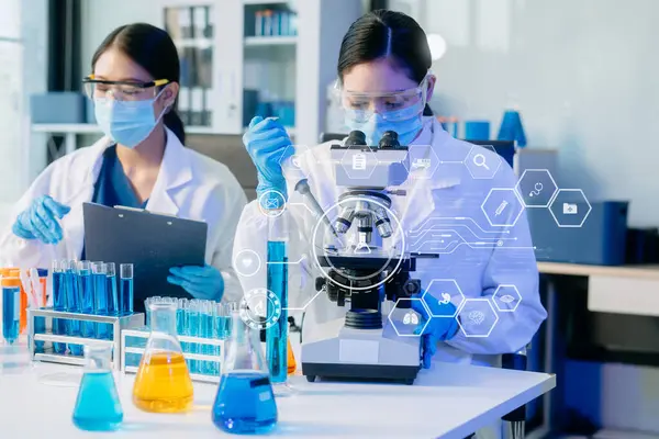 Yüz maskeli iki Asyalı kadın bilim adamı yüksek teknolojili bir tıp laboratuarında mikro pipetler kullanarak örnekleri analiz ederek biyokimyasal araştırma yapıyorlar. Tıbbi simgeleri olan sanal ekran 