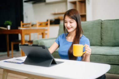 Modern evdeki kanepede otururken akıllı telefon ve tablet kullanan genç Asyalı kadın.