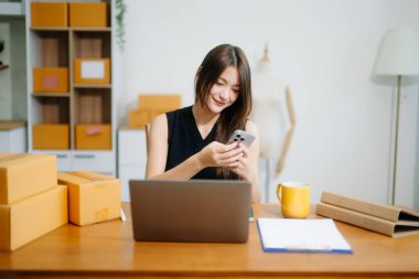 Startup small business SME, Entrepreneur owner woman using smartphone or tablet taking receive and checking online purchase shopping order to preparing pack product box.
