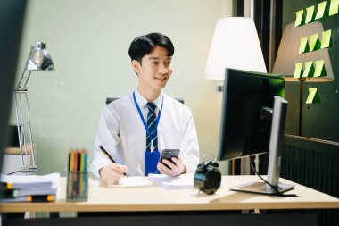 Ofiste çalışan genç Asyalı iş adamı. Ofisinde akıllı telefon ve bilgisayar var.