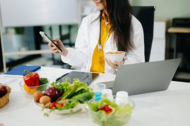 Akıllı telefon ve dizüstü bilgisayar kullanan Asyalı kadın beslenme uzmanı, klinik beslenme ortamında diyet planlaması, sağlık ve sağlık hizmetleri için taze sebze ve takviyeleri gözden geçirir..