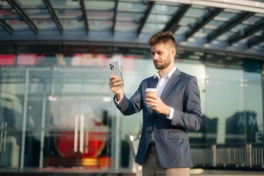 Genç iş adamı lider girişimci, profesyonel yönetici akıllı telefon ve kahve bardağını iş merkezinin arka planında büyük şehirde tutuyor..