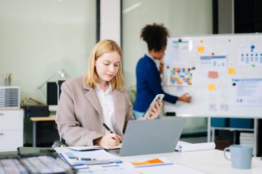 Kendine güvenen iş uzmanı, gülümseyen, dizüstü bilgisayarda yazan ve dijital tableti yaratıcı ofiste tutan çekici genç bir kadın..