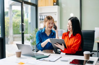 Asyalı iş arkadaşları dizüstü bilgisayar ve tablet kullanarak açıklama yapıyor. Takım çalışması, finansal pazarlama ekibi, modern ofis odasında otururken. 