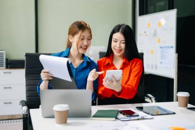 Asyalı iş arkadaşları dizüstü bilgisayar ve tablet kullanarak açıklama yapıyor. Takım çalışması, finansal pazarlama ekibi, modern ofis odasında otururken. 