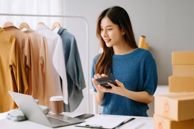 Startup small business SME, Entrepreneur owner woman using smartphone or tablet taking receive and checking online purchase shopping order to preparing pack product box.
