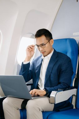 Çekici Asyalı erkek uçak yolcusu dizüstü bilgisayarla çalışırken rahat koltukta oturuyor. Stil içinde seyahat et, zarafetle çalış.