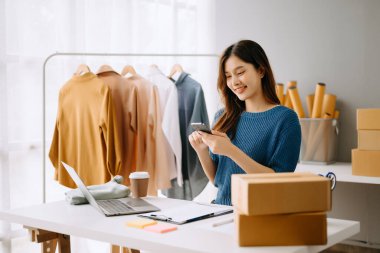Startup small business SME, Entrepreneur owner woman using smartphone or tablet taking receive and checking online purchase shopping order to preparing pack product box.