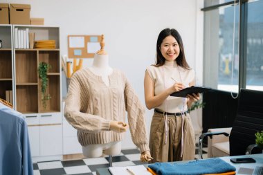 Asyalı terzi kadın terzi atölyesinde giysiler üzerinde çalışıyor. Güzel genç bayan moda tasarımcısı gülümsemesi ve stüdyo başarısından sonra