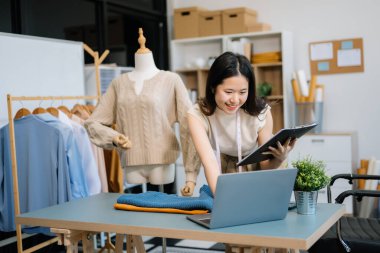 Asyalı terzi kadın terzi atölyesinde giysiler üzerinde çalışıyor. Güzel genç bayan moda tasarımcısı gülümsemesi ve stüdyo başarısından sonra