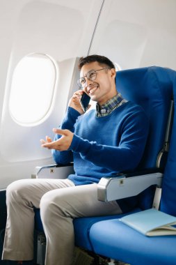 Asyalı bir adam uçakta bir koltukta oturuyor ve akıllı telefondan konuşuyor.