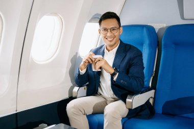 Asyalı işadamı uçak koltuğunda kahve içiyor ve kameraya bakıyor. 