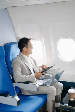 Asyalı işadamı bir uçak koltuğunda oturuyor ve laptopla tablet üzerinde çalışıyor. Seyahate çıkıyor..