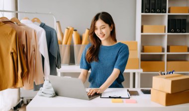 Asyalı terzi kadın terzi atölyesinde giysiler üzerinde çalışıyor. Güzel genç bayan moda tasarımcısı gülümsemesi ve Studi 'deki başarıdan sonra