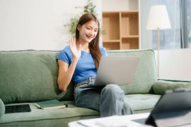 Genç Asyalı kadın modern evdeki koltukta otururken akıllı telefon ve tablet kullanıyor.