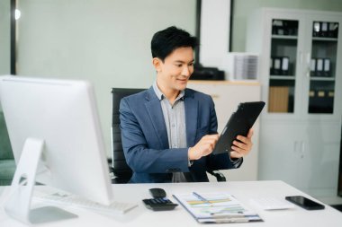 Çalışan not defteri, tablet ve dizüstü bilgisayarla çalışan genç Asyalı işadamı akıllı telefon, tablet ve dizüstü bilgisayarla konuşuyor.