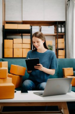 Genç işletme sahibi kadın paket kutuları hazırlayın ve tabletteki müşteriye teslimat için internetten siparişleri kontrol edin. İnternetten alışveriş konsepti. Evdeki kanepede. 