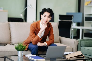 Asyalı girişimci işine odaklanmış, şık bir ofiste koltukta dizüstü bilgisayarla belgeleri analiz ediyor. İş için mükemmel, verimlilik için.