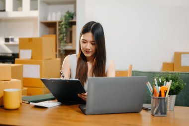 Başlangıçta küçük bir işletme olan KME girişimcisi kadın, ürün kutularını paketlemeye hazırlarken çevrimiçi satın alma siparişleri almak ve kontrol etmek için dizüstü bilgisayar kullanır  