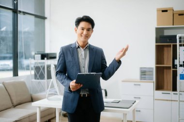 Genç yakışıklı Asyalı adam gülümsüyor. Not defteri, tablet ve dizüstü bilgisayarla yazmayı düşünüyor. Evden çalışıyor. Ofiste kameraya bakıyor. 