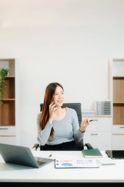 Genç ve çekici Asyalı iş kadını ofiste dizüstü bilgisayar ve telefonla çalışıyor.. 