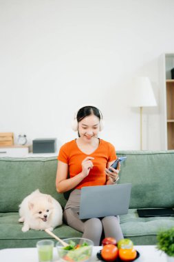 Parlak modern oturma odasında köpeğiyle koltukta dinlenen bir kadın dizüstü bilgisayar ve akıllı telefon kullanıyor.