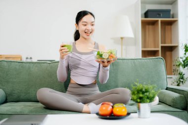 Spor yaptıktan sonra salata yiyip kanepede meyve suyu içen genç bir kadın. Aktif sağlıklı yaşam tarzı, temiz yeme konsepti